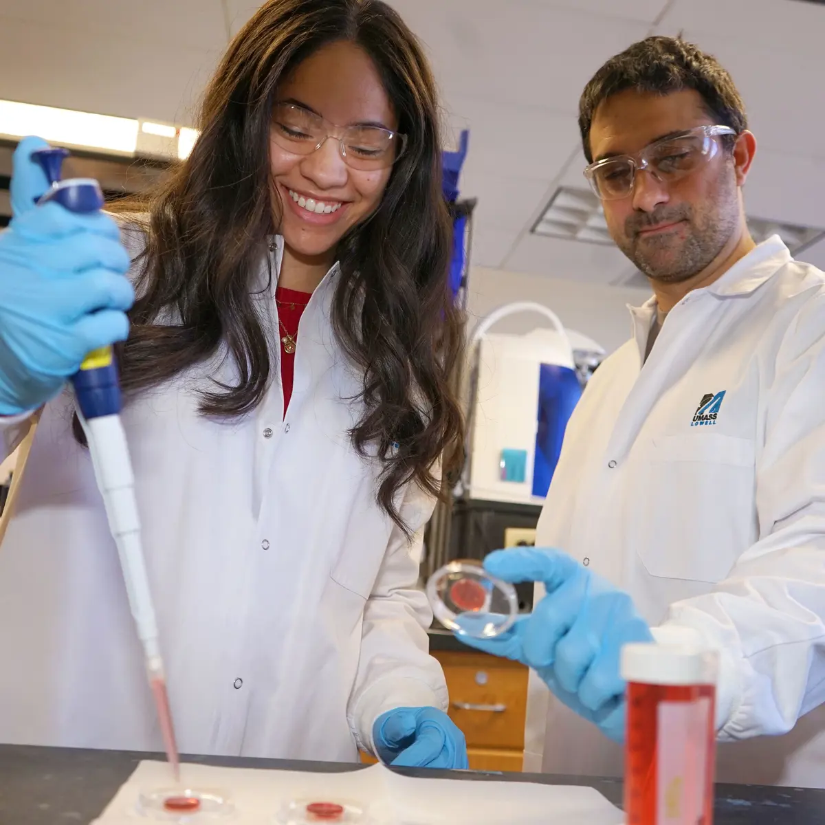 A student in a lab coat pipettes a specimin while another student in a lab coat holds up a petri dish.