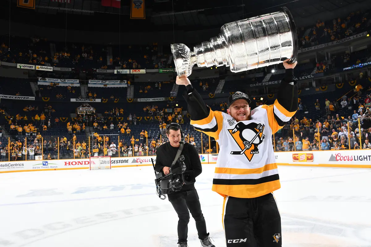 Chad Ruhwedel holding the Stanley Cup above his head on a hockey rink with a cameraman and a crowd behind him.