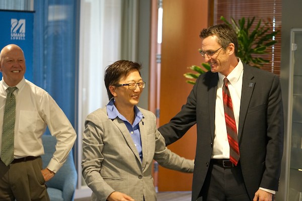 A smiling woman in glasses and a man in a suit greet each other while another man looks on
