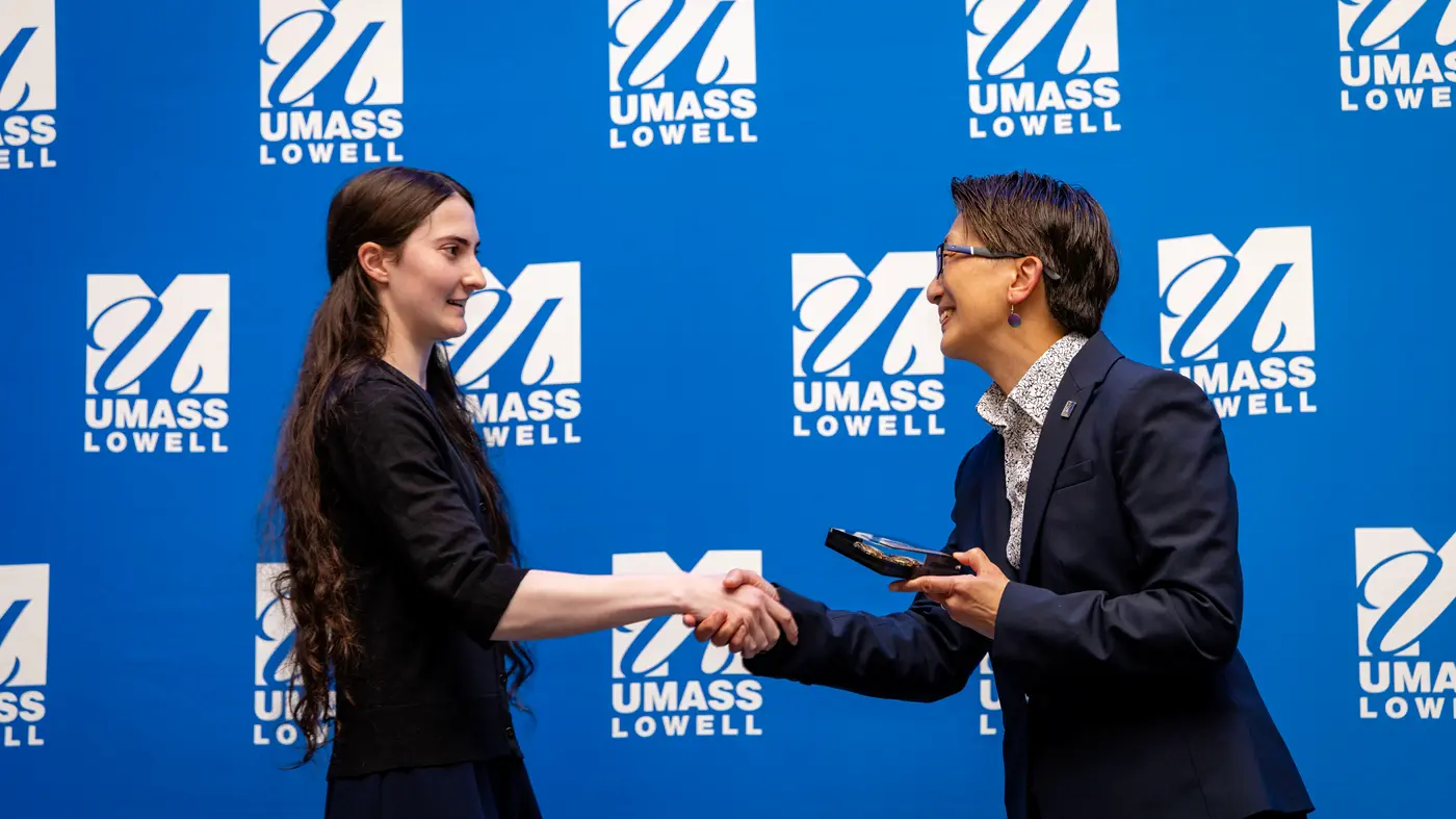 Chancellor Julie Chen shakes hands with a young woman whom she's presenting with an award in front of a UMass Lowell banner