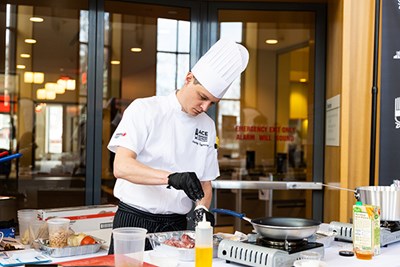 A chef in a white hat sprinkles salt on meat while cooking in a competition 