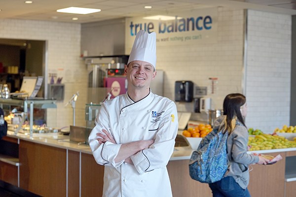 A man in a white chef's outfit poses for a photo with his arms folded while standing in a university dining hall