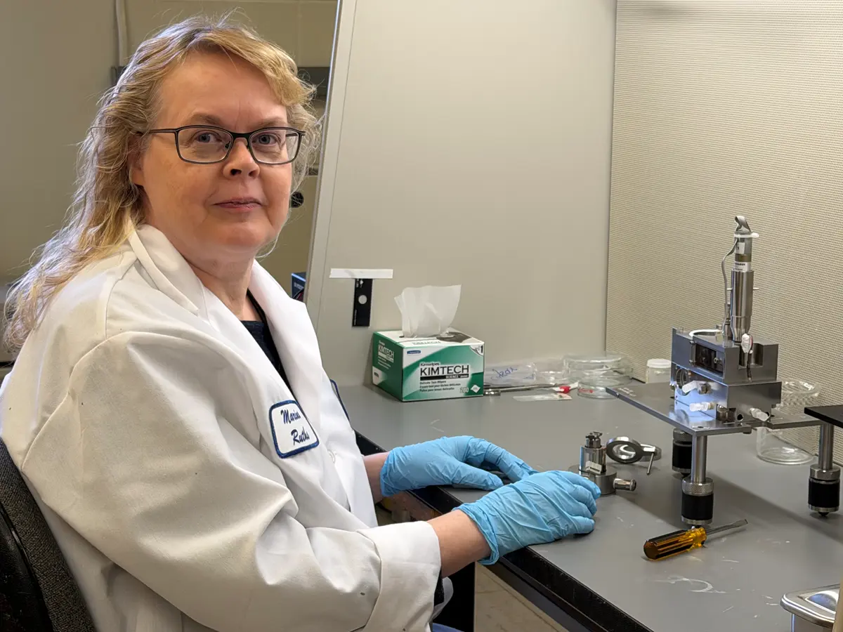 A woman in glasses and a white lab coat poses for a photo at a workbench.