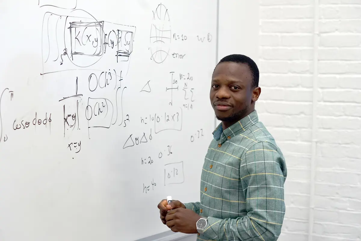 A young man poses for a photo while standing at a whiteboard and holding a marker.