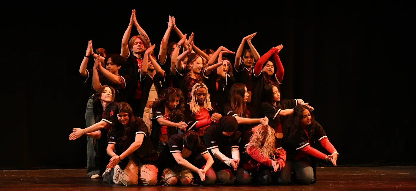 A diverse group of performers wearing matching black and red outfits in formation on stage.
