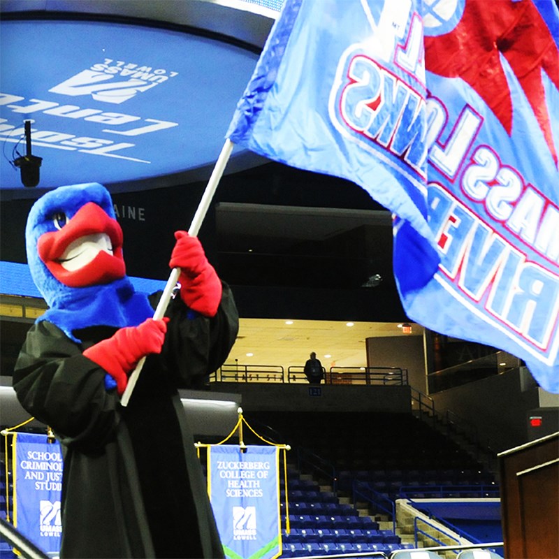 Mascot Rowdy the River Hawk wearing a black gown and waving a UMass Lowell flag