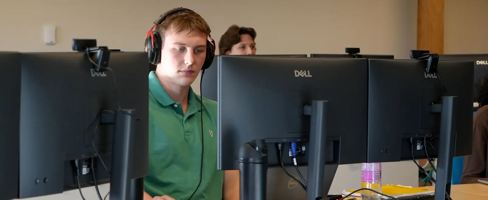 CyberTrust intern, Ryan Greaney, wearing headphones sitting at a Dell computer workstation with another person in the row behind.