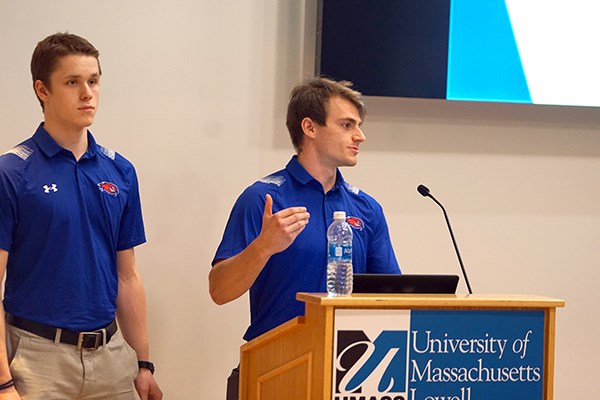A young man gestures while speaking at a podium while another young man looks on
