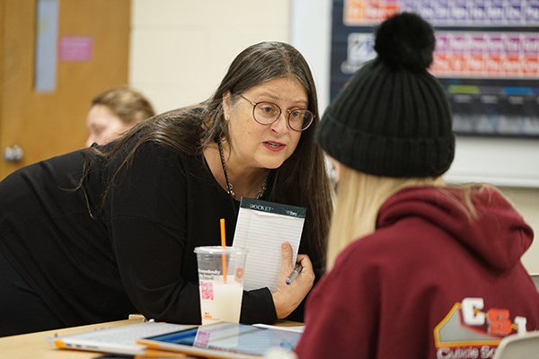 A professor in glasses leans on a table while talking to a female student wearing a black beanie