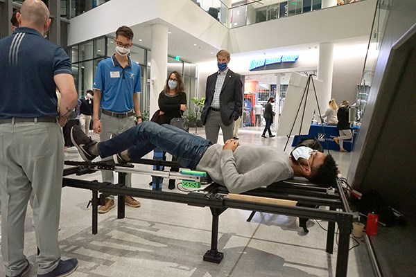 A man lies on a medical device table as a group of people stand around him inside a building