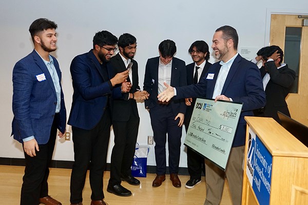 A group of students in suits accept a giant check from a man who is smiling