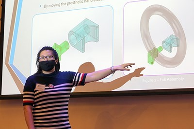A woman wearing glasses and a face covering points to a projector screen while making a presentation