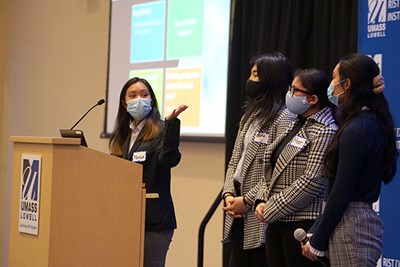 A woman in a face covering gestures while three women look on as they make a presentation