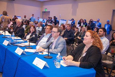 A panel of five judges sits at a table with a blue table cloth in a roomful of people