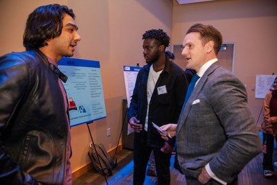 A man in a suit talks to a student next to their poster presentation while another man looks on
