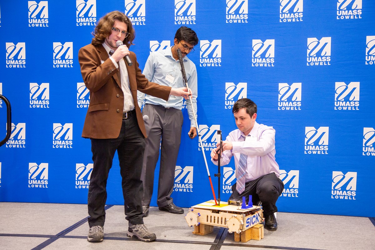 Three people demonstrate a robot that is attached to a walking cane on a stage.