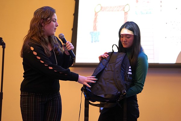 A woman holds a microphone and speaks while another woman holds a backpack during a presentation