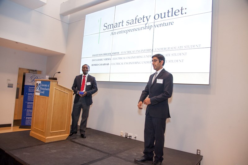 2 people in suits and ties presenting on a stage with a large screen behind them with words including: Smart Safety Outlet.