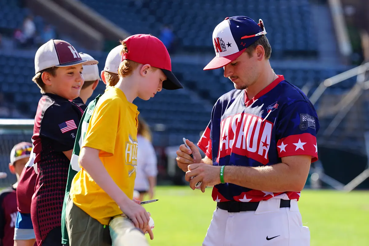 A college baseball player in a red, white and blue uniform signs an autograph for a group of kids along a fence.