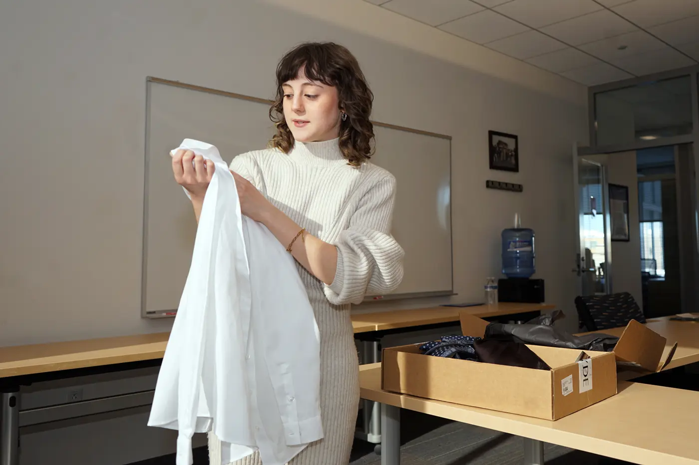 A young woman looks at a white blouse that she is holding while standing in a conference room.
