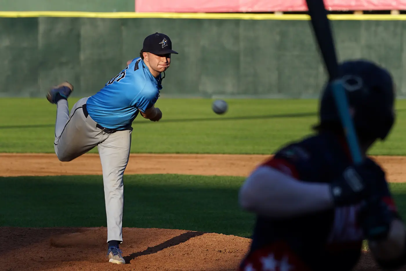 A baseball pitcher in a blue jersey delivers a pitch to a batter.