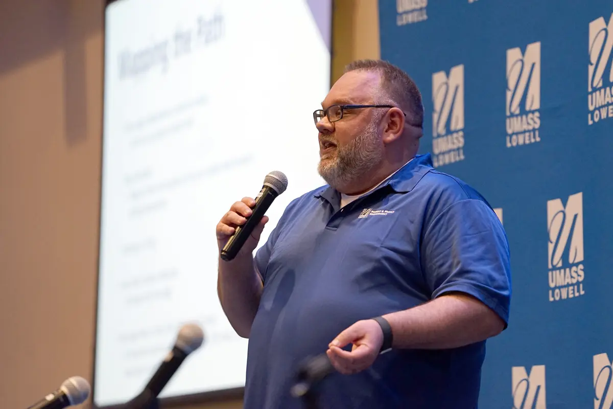 A man in a blue polo shirt and glasses holds a microphone while speaking on a stage.