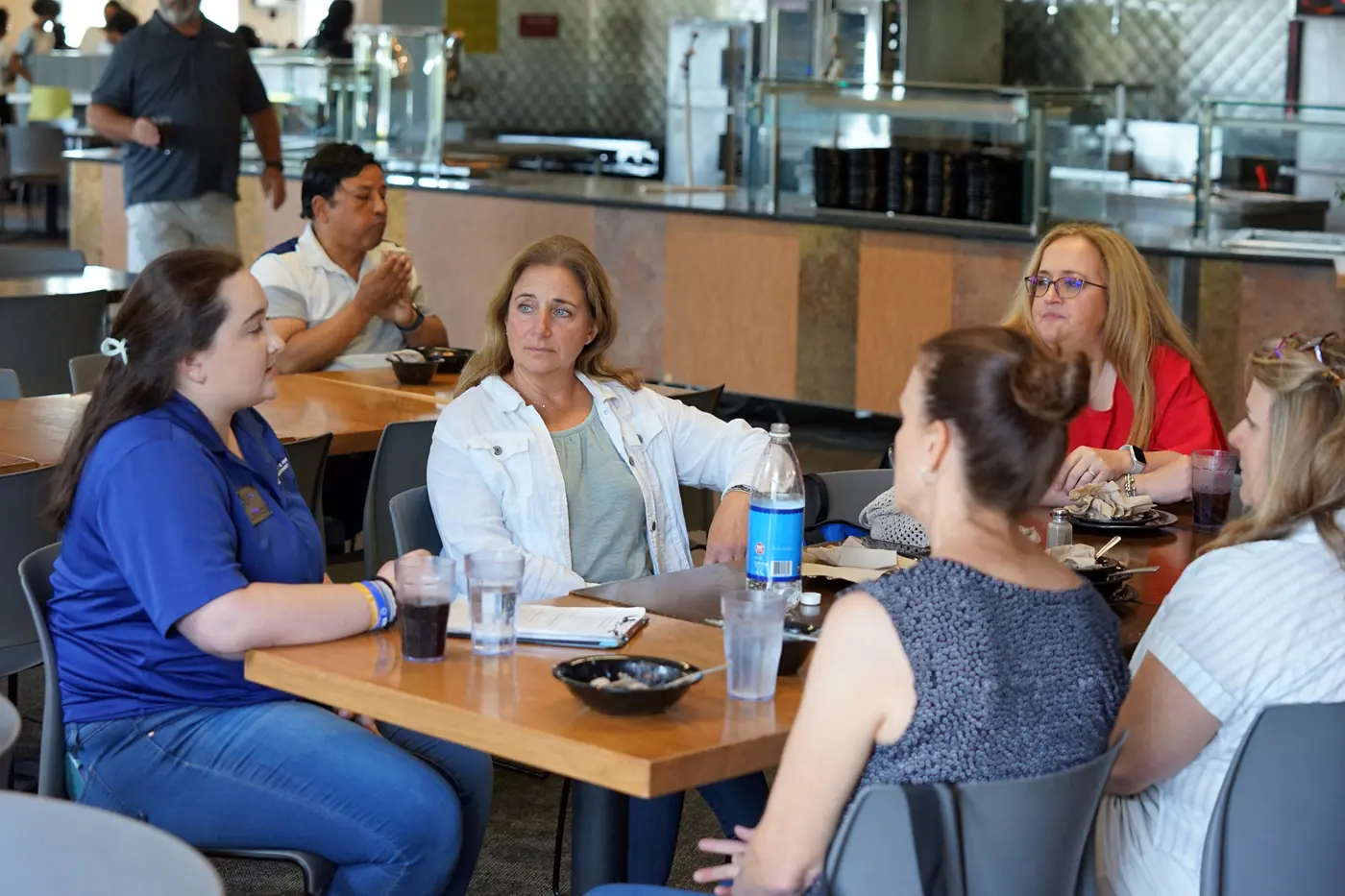 A student orientation leader in a blue shirt talks to four women seated at a dining hall table.