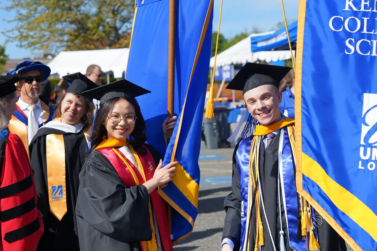 A young woman and man in college graduation gowns pose for a photo while holding blue and yellow banners outside.