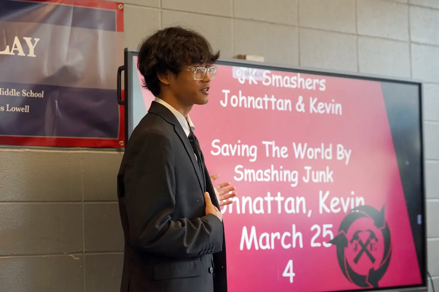 A person in glasses and suit makes a presentation in front of a screen with a red slide and text.