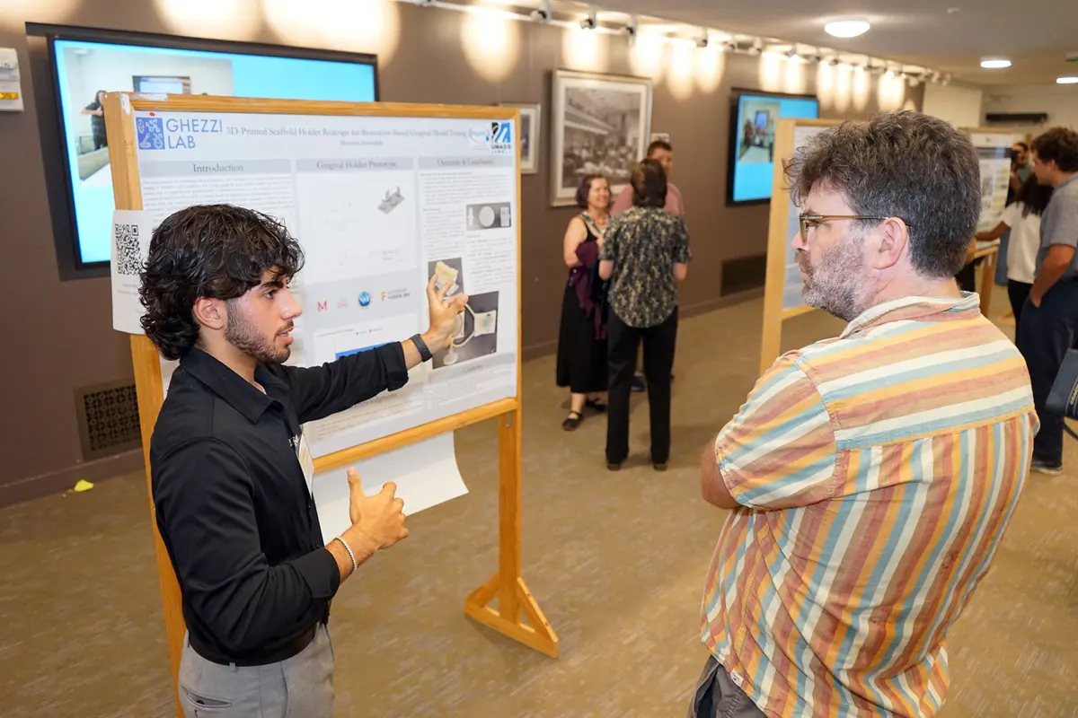 A young man with dark hair in a dark dress shirt gestures to a poster board while taking to a man in a colorful striped shirt.