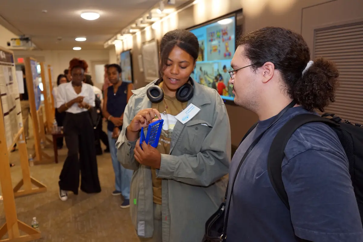 A young woman with headphones around her neck shows a piece of blue plastic to a young man with dark hair.