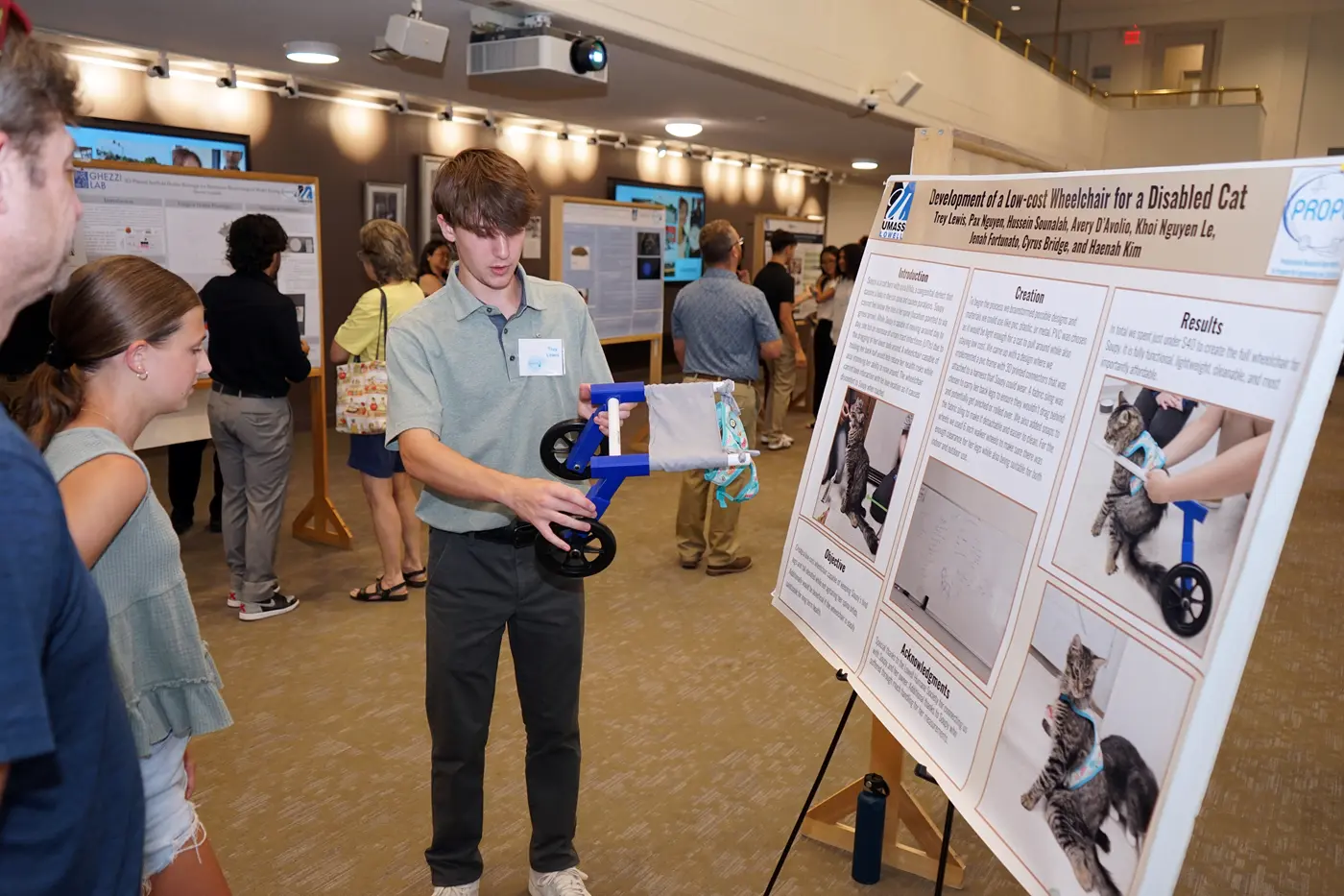 A young man shows a cat wheelchair that he helped build to a man and young girl at a poster session.