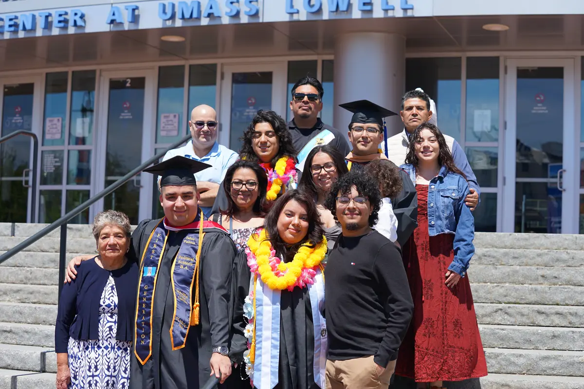 Four college graduates pose for a photo with family members on the front steps of an arena.