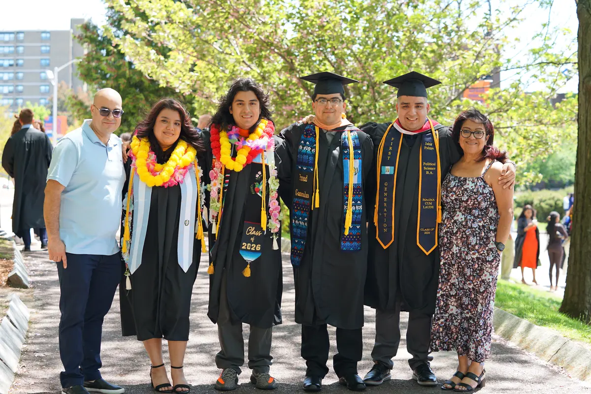 Four college graduates pose for a photo with their parents on sidewalk outside.