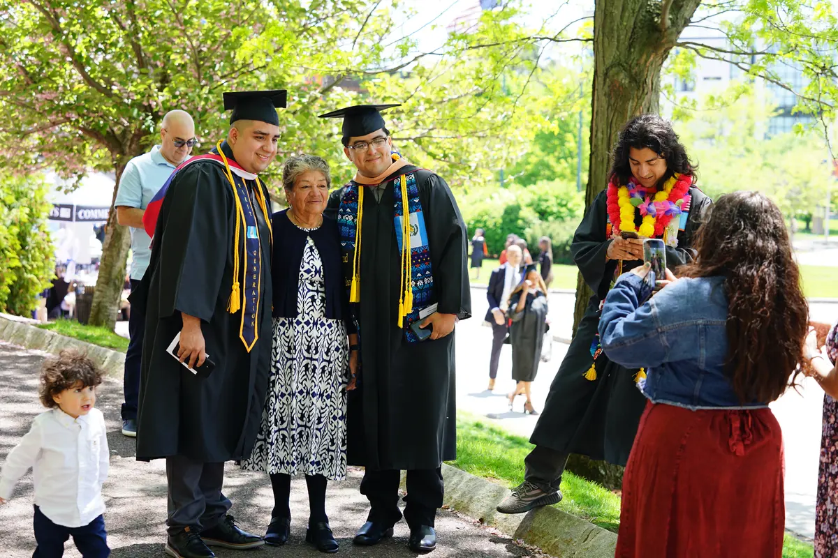 Two college graduates have their photo taken with their grandmother while a young child walks past.
