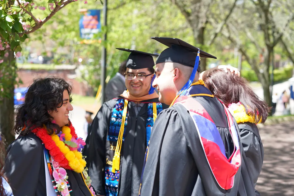 Four college graduates stand in a circle and chat outside on a sunny day.