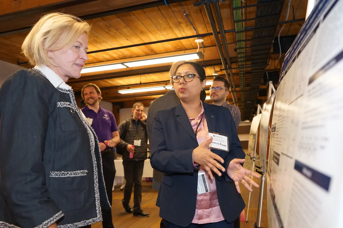 A woman in glasses gestures with her hands while she presents a poster to a woman with short blonde hair. 