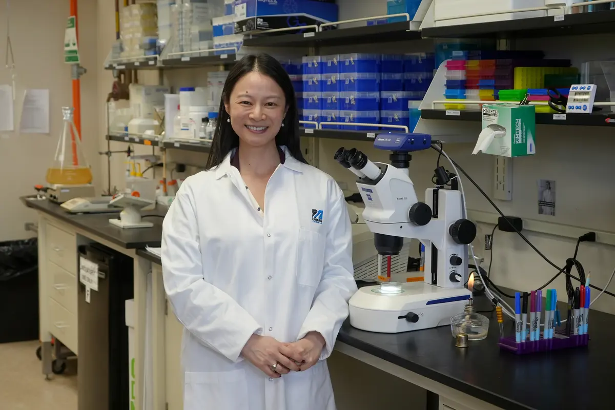 A woman with dark hair is wearing a lab coat and standing next to a microscope on a bench in a lab.