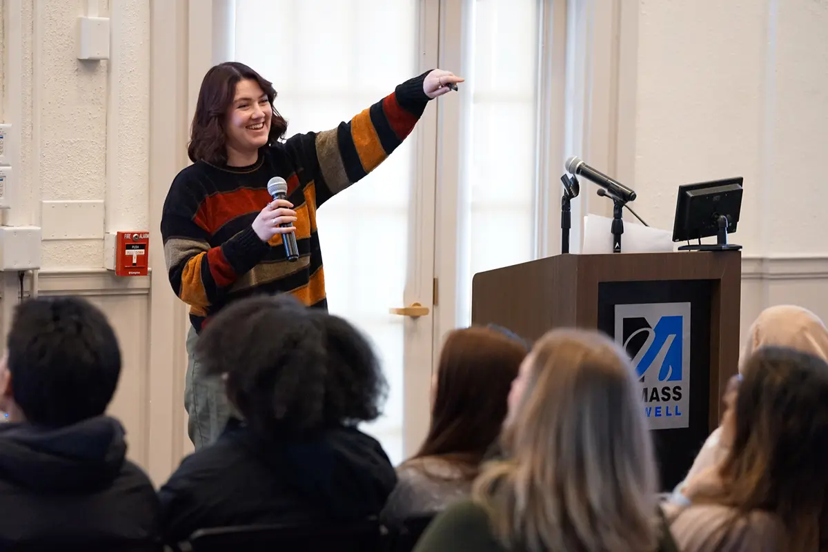 A young woman smiles while holding a microphone and pointing while talking to a group of students.