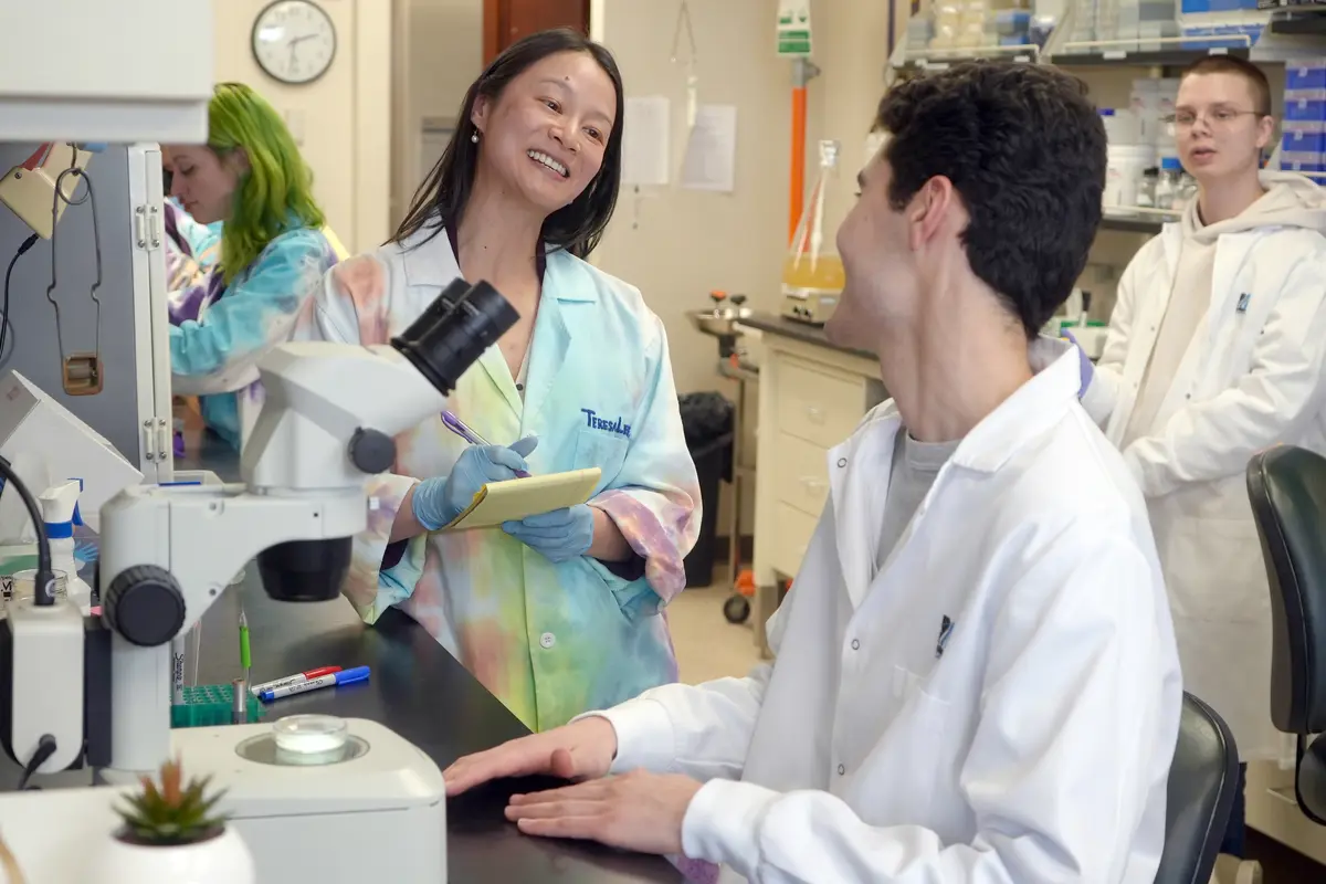 A woman in a lab coat holds a writing pad and smiles at a young man who is seated at a microscope in a lab.