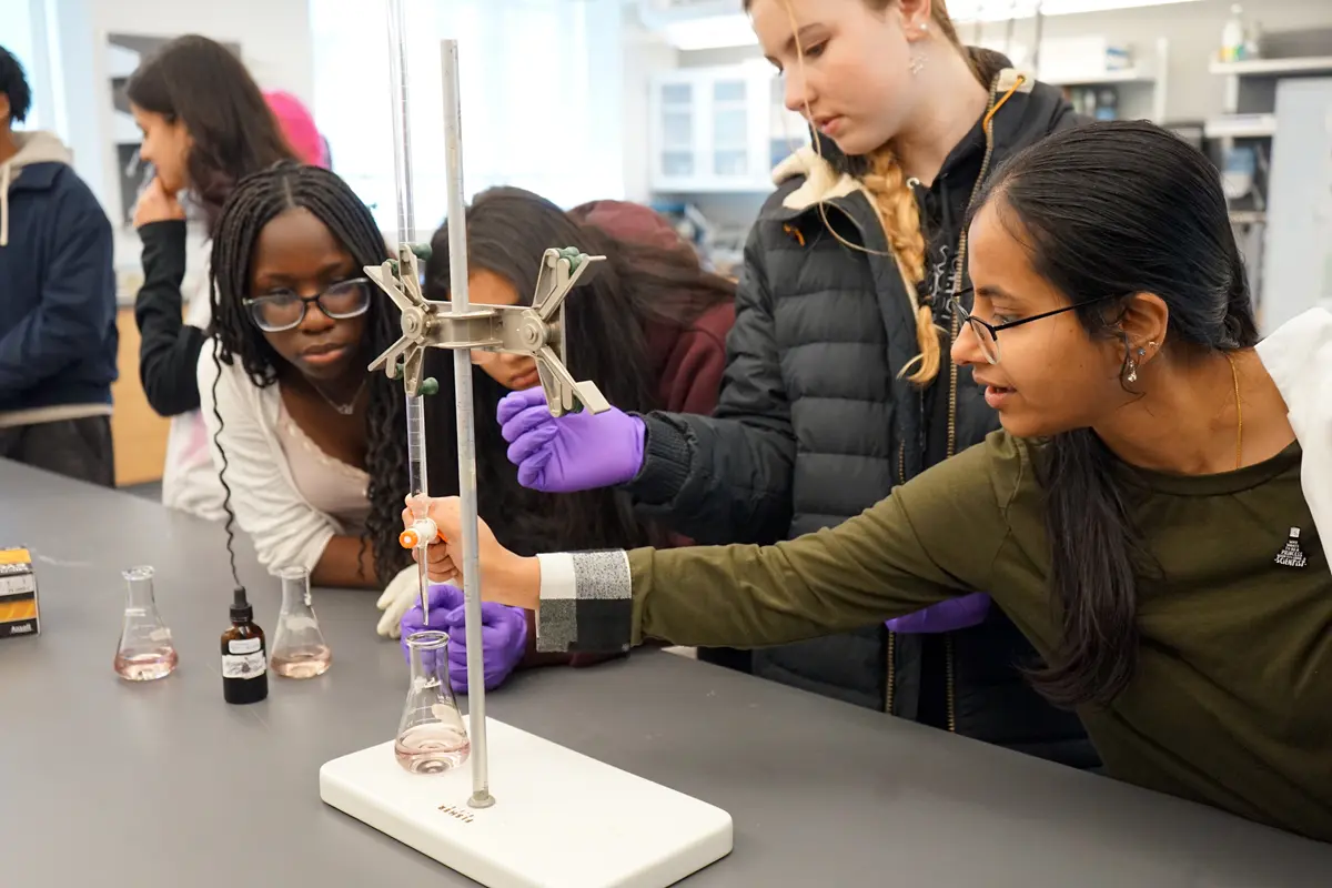 A young woman points to a research poster while talking to two high school students in a large room.