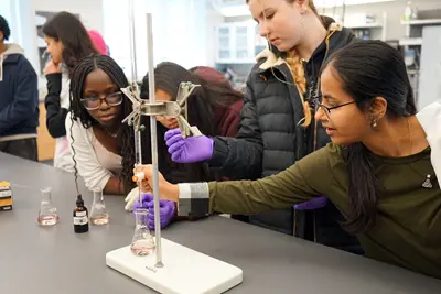 A young woman points to a research poster while talking to two high school students in a large room.