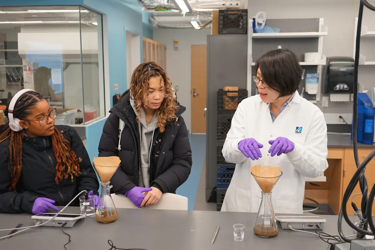 A professor with dark hair and wearing a lab coat talks to two high school students while standing in front of a glass carafe with a filter on it.