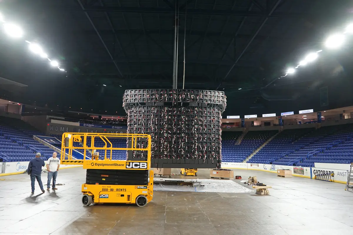 Two men talk while standing next to a video scoreboard that is under construction in an arena.