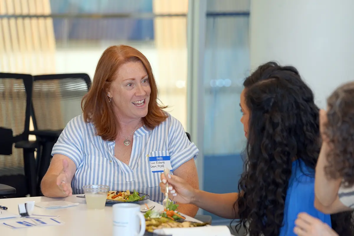 A woman in a while and blue striped shirt gestures while talking to another woman while eating lunch in a conference room.