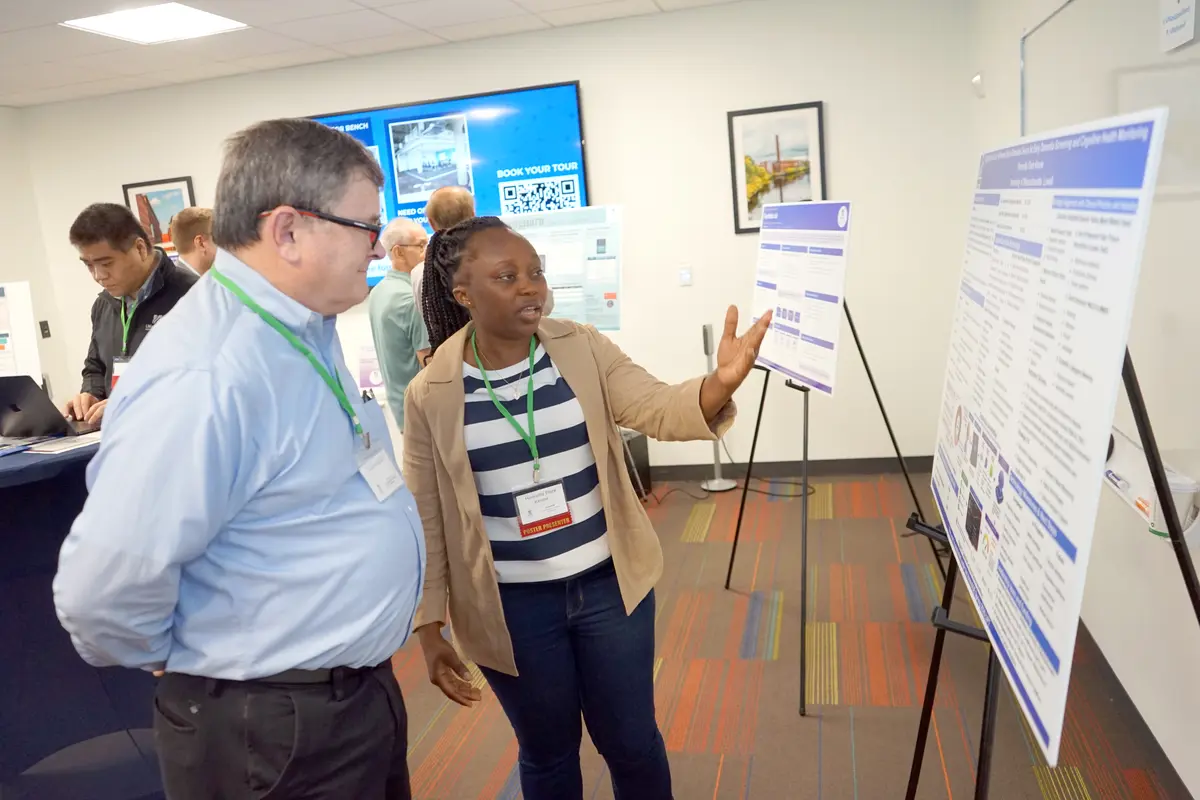 A woman with a striped shirt gestures toward a poster on an easle while talking to a man in a room.