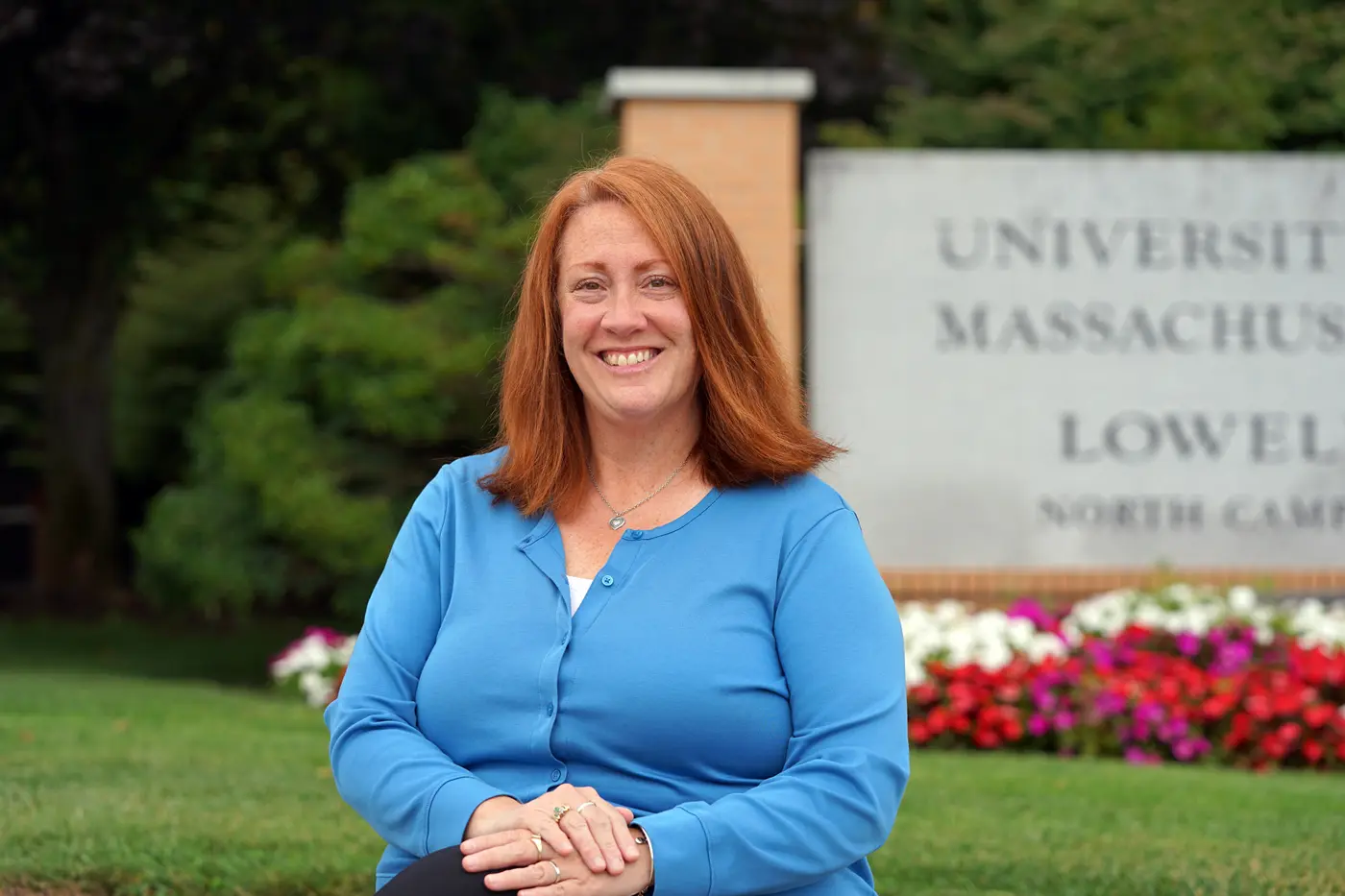 A woman with red hair and wearing a blue sweater poses for a photo outside in front of a college campus sign.