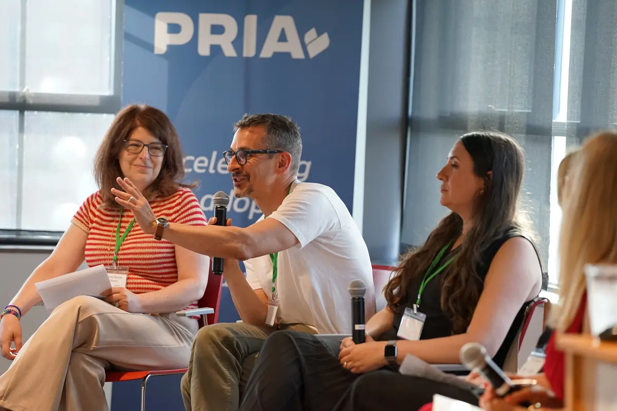 A man in a white T-shirt and glasses gestures while speaking into a microphone and seated between two women.