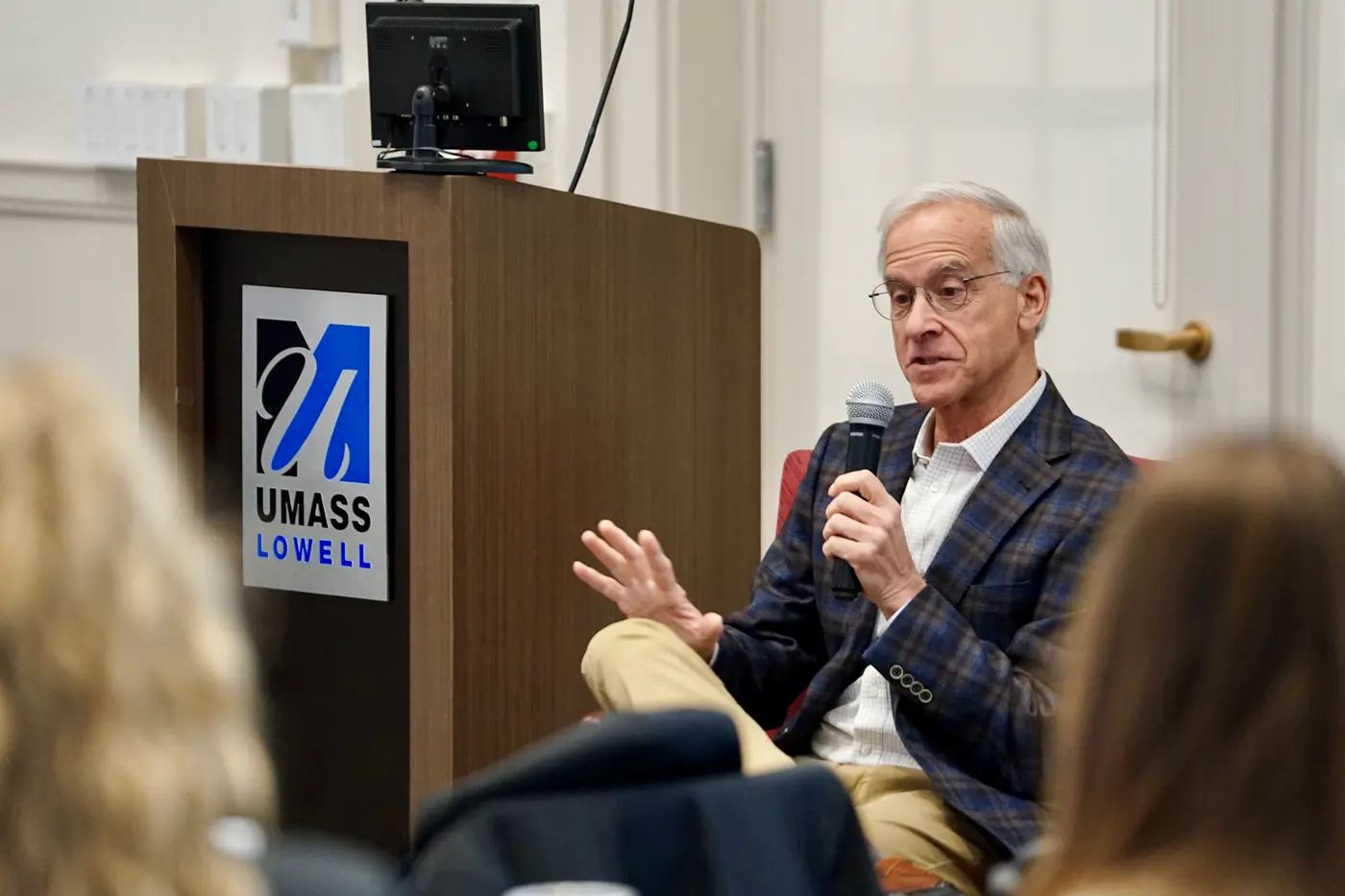 A man wearing glasses and a blazer holds a microphone and talks to students while seated in a chair.