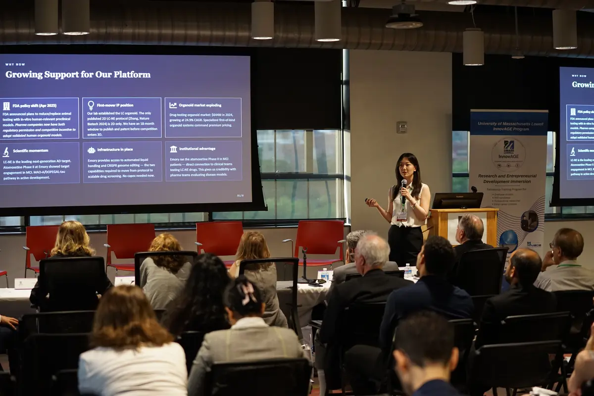 A young woman with long dark hair gives a presentation to people sitting in chairs.